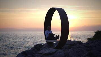 A person is working on their laptop while enjoying the beautiful sunset near the ocean video