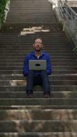 A Man Engaged in Work on His Laptop While Sitting Comfortably on a Staircase Outside video