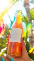 A hand holding a glass bottle of fresh tropical juice with a blank label, set against a bright, colorful background with palm leaves and sunlight photo