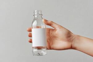 A clean mockup of a transparent plastic bottle with a blank white label held by a hand against a neutral gray background photo