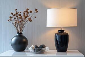 vase with dried flowers, glass bowl with sea stones and table lamp on white surface photo