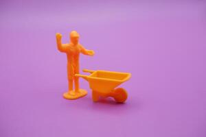 Mini Workers Raising Hands with Yellow Wheelbarrow Isolated on Purple Backdrop photo