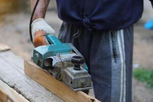 Closeup carpenter using electric planer with wooden plank in carpentry worksite. Concept, tool or equipment for construction work. Using with careful and skillful for safety. photo