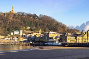 Sandy beach with waterfront buildings and hills in the background at sunset in San Sebastian, Spain. Coastal cityscape and natural landscape photo