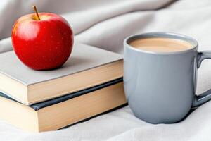 Cozy reading corner features a stack of books with beige covers, a gray ceramic mug of coffee, and a vibrant red apple on a soft linen tablecloth photo