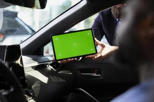 Dealership salesman showing vehicle make, model and features during test drive using mockup tablet. Showroom agent helps customer evaluate automobile, showing green screen device photo