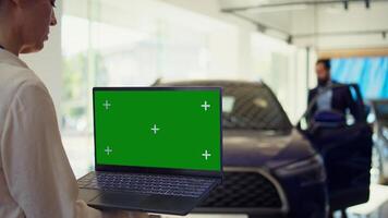 Customer in showroom looking for new vehicle to purchase or lease using green screen laptop. Woman evaluating automobiles in dealership before buying using chroma key notebook, camera B photo
