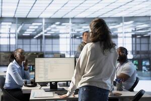 Manager supervising multiethnic team working in data center, raising from desk to make announcement. Team leader discussing with employees monitoring energy consumption across server rigs photo