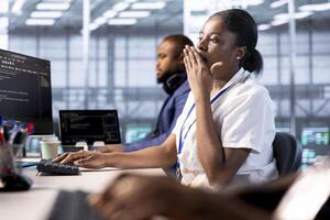 Bored data center technician yawning while creating technical documentation for software applications. Exhausted woman feeling tired after overworking in server hub maintaining databases photo