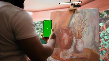 Young artist looking at isolated chroma key display in his painting atelier, using device next to canvas with colorful masterpiece. Painter creating rustic artwork for gallery exhibition. Camera B. photo