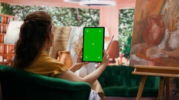 Painter holding a device with isolated mockup screen in her art studio, looking at masterpiece with oil paints on canvas. Beautiful rustic artwork for a fine art gallery, creativity. Camera B. photo