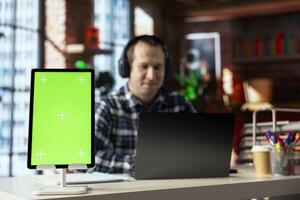 Close up of mockup tablet next to smiling employee at work watching videos on social media using laptop. Focus on green screen device next to employee using social network website on notebook photo