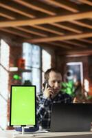 Focus on green screen tablet with home office worker in blurry background using laptop during phone call. Chroma key device next to man in apartment discussing with coworker in telephone call, closeup photo