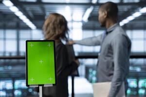 Isolated screen mockup tablet in front of engineers in blurry background working in server farm, analyzing data. Close up of chroma key tablet next to employees examining data center photo