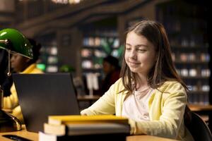 Primary school student sitting at desk in the campus library and writing on laptop, preparing notes and ideas for future lessons. Scholar girl using digital gadget for assignments. photo