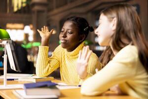 Two diverse schoolgirls attending an online class via call in the library, focused on distance learning and completing a school assignment using modern digital technology. Webinar work. photo