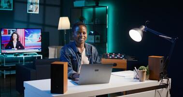 Portrait of smiling african american woman at desk typing on laptop keyboard, solving tasks. Joyful remote worker using notebook to send emails to business partners from living room, camera B photo