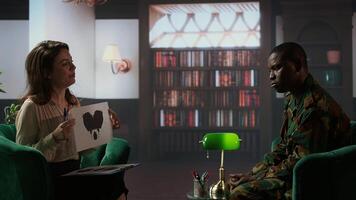 Counselor specialist presents a Rorschach inkblot test to an army trooper in military uniform during a psychoanalysis. Diverse people at therapy in a warm office with bookshelves. Camera A. photo