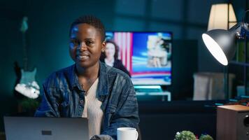 Portrait of smiling african american woman at desk typing on laptop keyboard, solving tasks. Joyful remote worker using notebook to send emails to business partners from living room, camera A photo