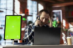 Close up of green screen tablet next to unhappy manager in telephone call with employee offering negative feedback. Focus on mockup device in front of supervisor berating worker during phone call photo