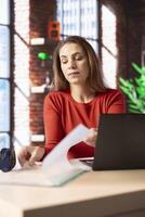 Freelancer does her research by reading articles and data driven papers at home, collecting information for a new project. Woman multitasking and studying files with handwritten notes at the desk. photo