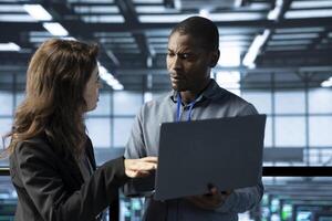 System administrators in data center using laptop, analyzing metrics, optimizing storage volumes. Server farm IT experts using notebook, talking, ensuring seamless operations photo
