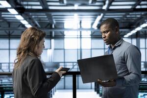 Computer scientists in data center using laptop and tablet, executing security patches, verifying firewall configurations. Man and woman performing equipment vulnerability scans in server farm photo