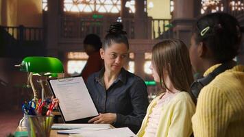 Female mentor providing instruction to diverse pupils during private lessons at the library, assisting with homework and school projects in the study area. Tutoring session for guidance. Camera A. photo