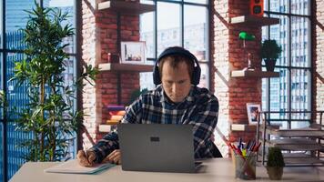 Worker listening music, analyzing data on laptop at desk, compiling analytical reports. Employee wearing headphones, using notebook device, writing down information, camera A photo