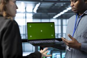 Teamworking coworkers in data center using automation tools on green screen mockup laptop. Engineers ensuring continuous uptime of server farm, managing data flows with chroma key device photo