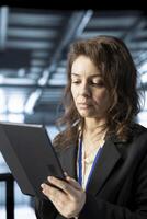 Technician in data center workspace using tablet, configuring network virtualization. Woman in server room updating storage arrays with device, verifying settings to maintain stable performance photo