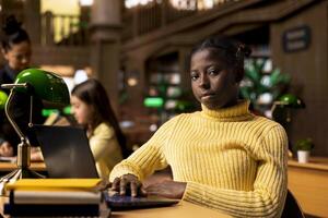 African american schoolgirl writing class notes at a study desk in a library, working hard on a school project with online information on laptop. Pupil preparing for lessons at the academic institute. photo