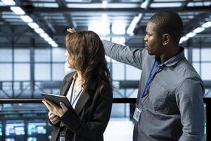 Coworkers holding tablet, looking around data center, managing server virtualization. Server room employees finetuning network protocols, enforcing advanced security measures photo