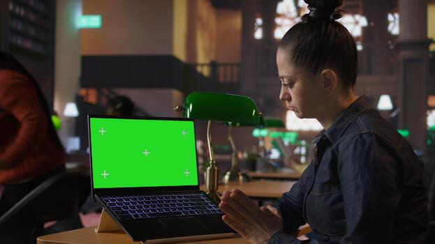 Professional librarian working on her laptop next to mockup screen in a public library, providing academic resources and guidance. Evaluating educational information within the database. Camera A. photo