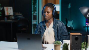 Remote worker with wireless headphones working from living room office, sending emails. Freelancer doing internet research using laptop while listening to music to make time pass faster, camera A photo