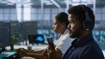 Worker in data center wearing headphones taking break from implementing AI systems, using smartphone. Focus on artificial intelligence server farm employee using mobile phone, camera A photo