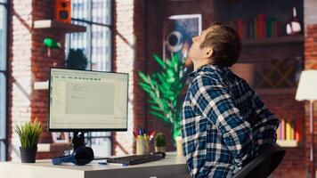 Software developer at computer coding, stretching joints after sitting at desk too long. IT expert in home office doing muscle exercises after feeling pain from incorrect posture, camera B photo