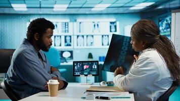Black male patient bringing x ray test results to medic for scan interpretation, reviewing the reports for diagnostics. Medic expert evaluating the radiography film in cabinet. Camera B. photo