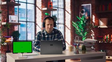 Green screen tablet next to employee completing spreadsheets on laptop and listening music. Chroma key device next to worker in home office wearing headphones editing documents, camera A photo