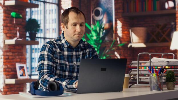Man seated at home office desk using laptop, answering phone call while checking emails displayed on screen. Worker in telephone call with friend at work while reading messages in inbox, camera B photo
