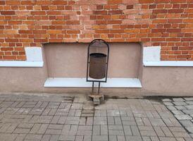 A garbage can on the background of a red brick facade and a pink plastered basement with a ventilation grille photo