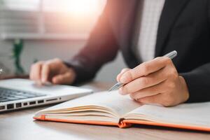 Close-up of a business professional writing in a notebook at a desk with a laptop, illuminated by soft sunlight. photo