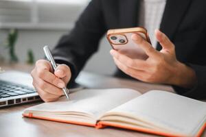 Business Professional Writing Notes While Using Smartphone at Office Desk with Laptop and Notebook photo