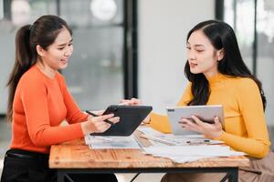 Two women in a modern office setting, discussing work using digital tablets and documents on a wooden table. photo