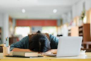 A tired student rests their head on a desk surrounded by a laptop and books in a well-lit classroom environment. photo