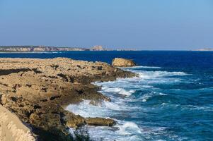 Waves crash against rocky coastal cliffs under a clear blue sky in Cyprus during a sunny day. photo