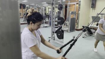A woman is using a rope machine at the gym to workout. A man stands nearby in the background. The gym has various workout machines. The scene is indoors. video
