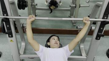 A woman performs a bench press using a weight machine in a gym. Another person is visible in the mirror. There are weights on the bar. The floor is green. video