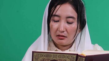 Close-up of a woman reading from an open book quran with Arabic scrip. She is wearing a white headscarf and is focused on the text. isolated on a green background or green screen video