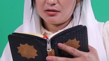 A person with a white veil is holding an open book, possibly the Quran, and is reading it, isolated on a green background or green screen video
